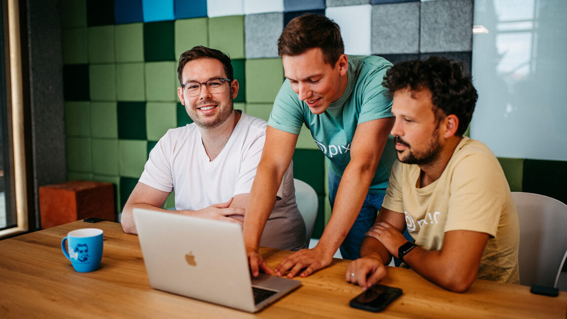 Drei Männer sitzen und stehen um einen Laptop mit der pixx.io DAM-Software auf einem Holztisch und arbeiten in einem modernen Büro mit einer grün-grau karierten Wand zusammen.