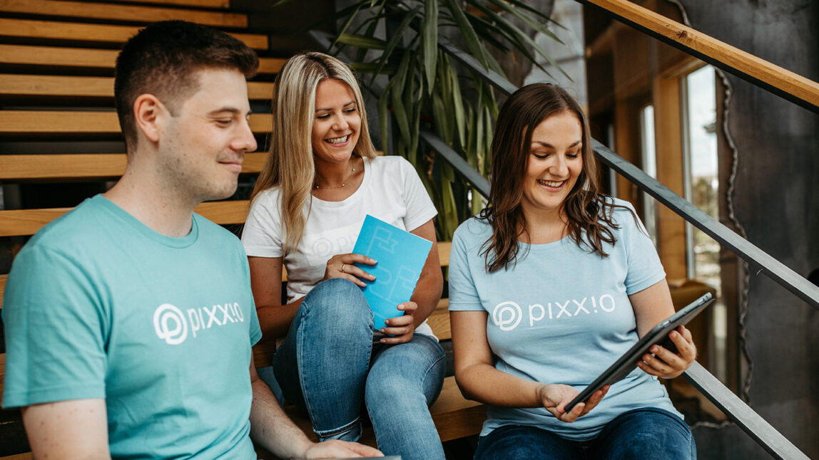 Drei Personen sitzen auf einer Holztreppe in einem Innenraum und tragen Marken-T-Shirts. Zwei Frauen halten ein Tablet und ein Notebook, auf dem pixx.io abgebildet ist, während der Mann zusieht. Sie scheinen eine lockere Diskussion über DAM-Lösungen zu führen.