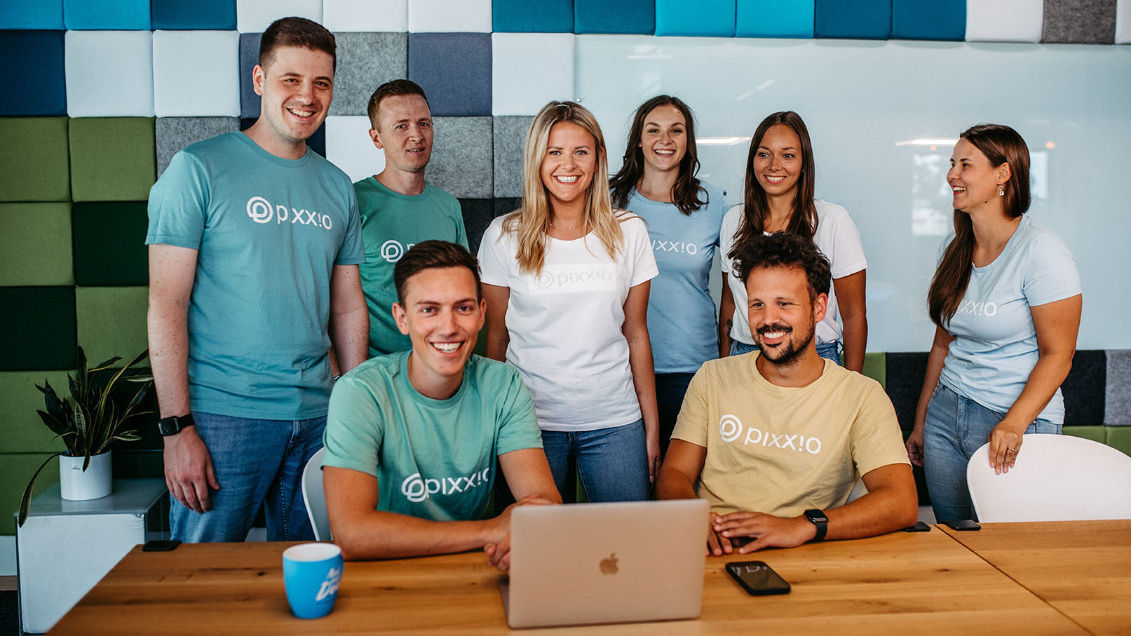 Eine Gruppe von acht Personen mit "pixx.io"-Shirts posiert und lächelt um einen Laptop herum an einem Holztisch in einem modernen Büro und hebt ihre Leidenschaft für DAM-Lösungen vor einer bunten Wand hervor.