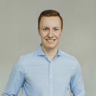 Ein junger Mann mit kurzen roten Haaren und einem hellblauen Button-up-Hemd steht vor einem schlichten hellen Hintergrund und lächelt in die Kamera.