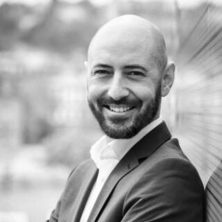 Bald man with a beard wearing a suit, smiling and standing with arms crossed outdoors near a blurred background.