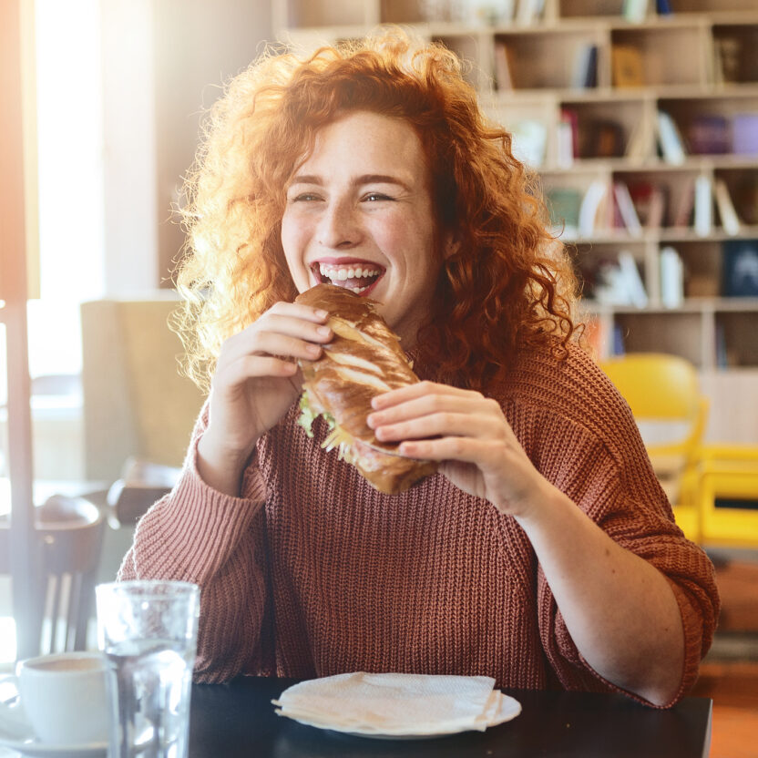 Frau mit lockigem rotem Haar, die einen rosa Pullover trägt, lächelt, während sie nach einem optimierten Einkauf ein Sandwich in einem Café genießt. Ein Glas Wasser und eine Serviette stehen auf dem Tisch, im Hintergrund stehen Bücherregale und Stühle.