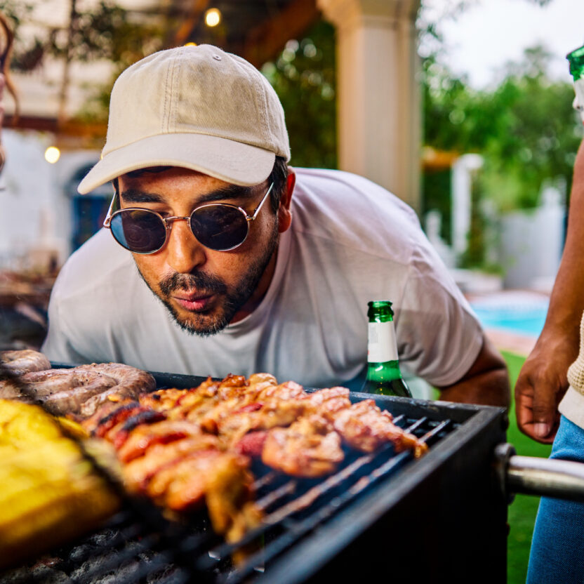 Ein Mann mit Sonnenbrille und Mütze pustet auf das Essen, das auf einem Grill zubereitet wird, und hält eine Bierflasche in der Hand, während er mit Freunden bei einem Treffen im Freien über die Optimierung der Lieferkette diskutiert.