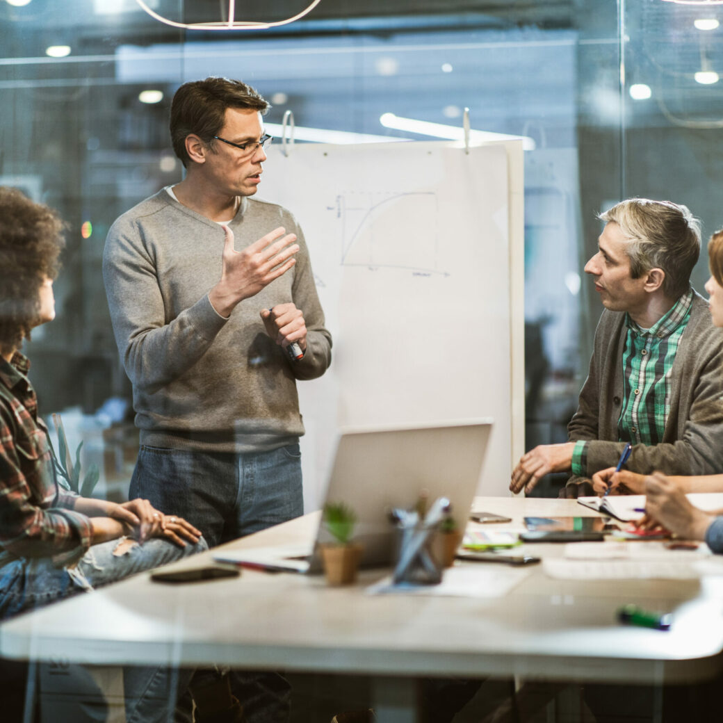 Ein Mann steht und präsentiert fünf sitzenden Kollegen in einem modernen Konferenzraum eine Grafik auf einem Flipchart zum Thema nachhaltige Beschaffung.