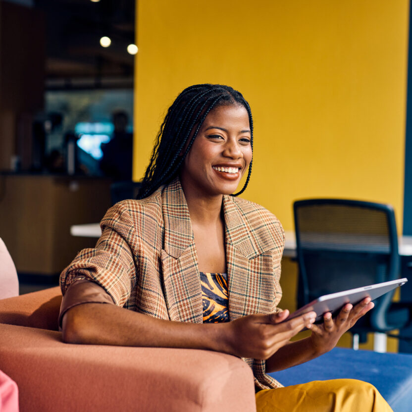 Eine Frau sitzt auf einer Couch, hält ein Tablet in der Hand und lächelt. Neben ihr sitzt ein Mann in einer modernen, farbenfrohen Büroumgebung, die ihr Engagement für nachhaltige Beschaffung widerspiegelt.