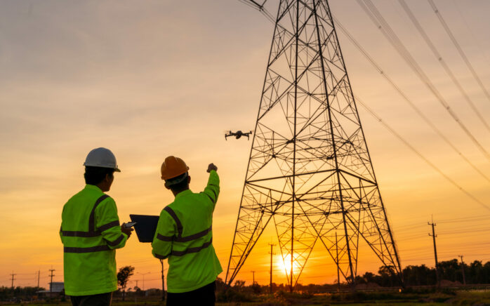 Two workers in safety gear observe and control a drone inspecting an electrical transmission tower at sunset.