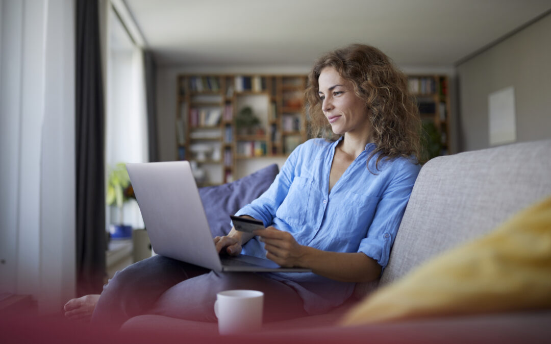 Eine Frau sitzt auf einem Sofa mit einem Laptop und einem Smartphone in der Hand, vor ihr eine Kaffeetasse, hinter ihr ein Bücherregal - vertieft in Aufgaben des Product Experience Management.