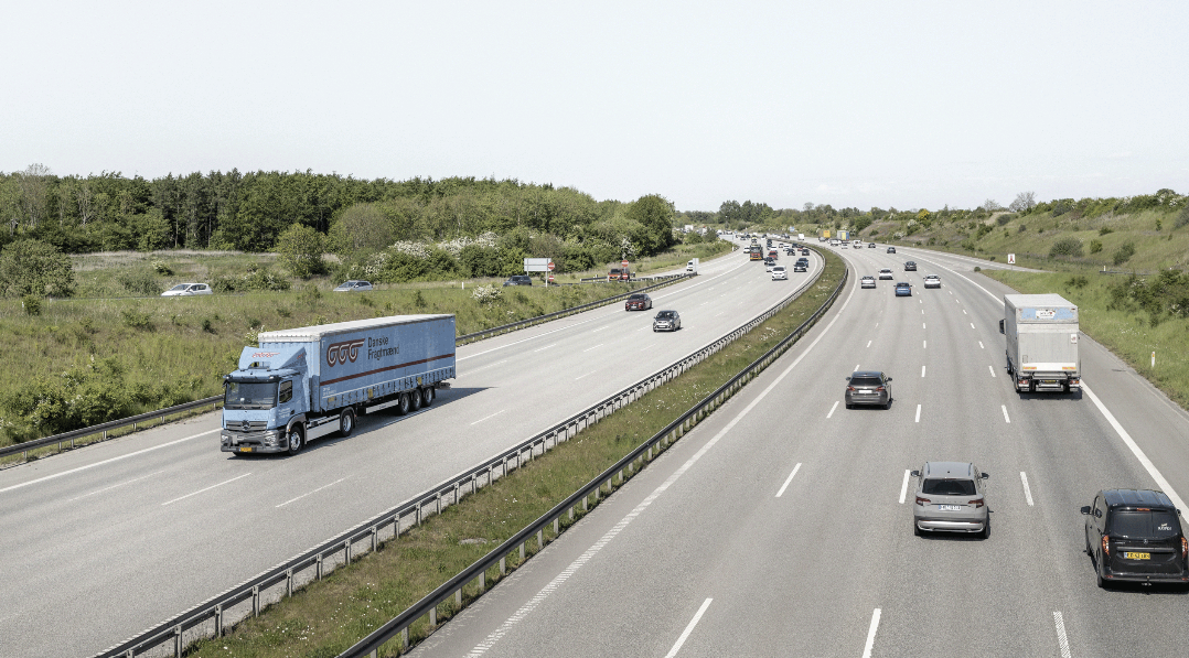 Eine Autobahn mit mehreren Fahrspuren in jeder Richtung, auf der Autos und Lastwagen auf beiden Seiten fahren, umrahmt von Grünflächen und Bäumen.