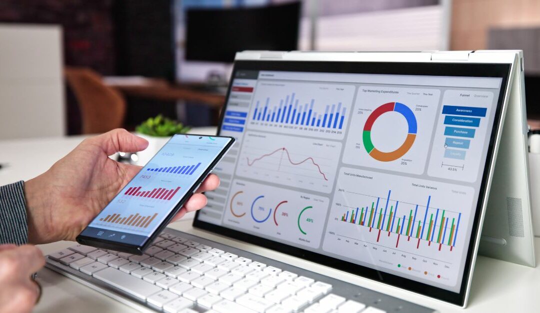 A person is holding a smartphone displaying charts. Next to them is a computer monitor showing various colourful charts, graphs and data visualisations.