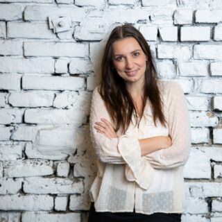 Eine Frau mit langen braunen Haaren und weißer Bluse steht mit verschränkten Armen lächelnd vor einer weiß gestrichenen Ziegelwand.