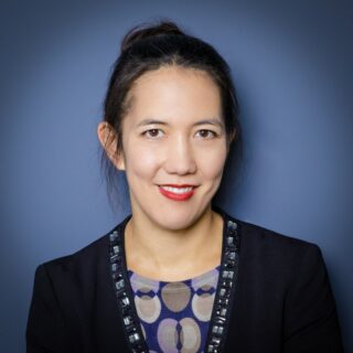 Eine Frau mit dunklem Haar in einem Dutt, die einen dunklen Blazer über einer gemusterten Bluse trägt, lächelt in die Kamera vor einem einfarbig blauen Hintergrund.