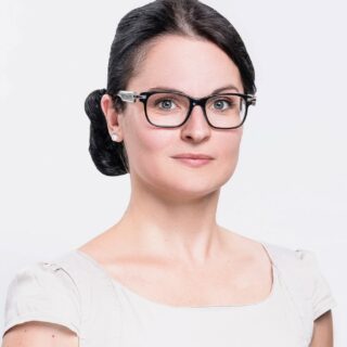 Eine Frau mit dunklen Haaren in einem tiefen Dutt, einer Brille und einem hellen Oberteil blickt direkt in die Kamera vor einem schlichten Hintergrund.