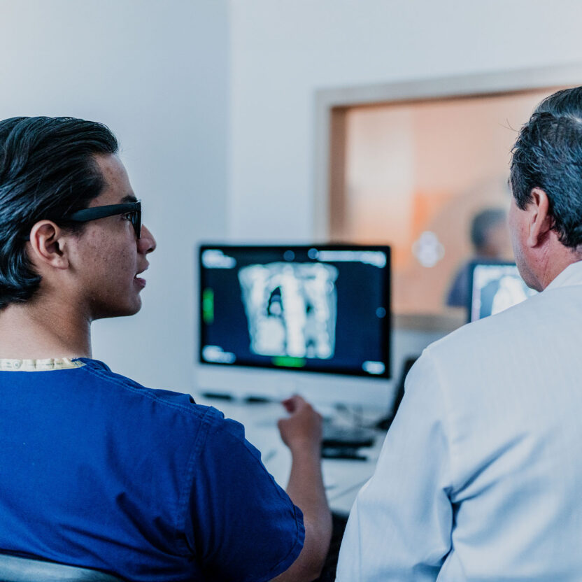 Zwei Personen in medizinischer Kleidung betrachten in einem klinischen Umfeld Diagnosebilder auf einem Computermonitor, wobei sie der Kamera den Rücken zuwenden.