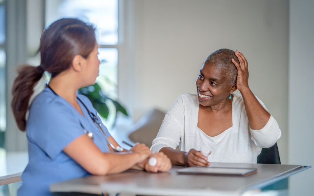 Eine medizinische Fachkraft in blauem Kittel unterhält sich mit einer lächelnden Frau in einem weißen Oberteil, die an einem Tisch in einem gut beleuchteten Raum sitzt.