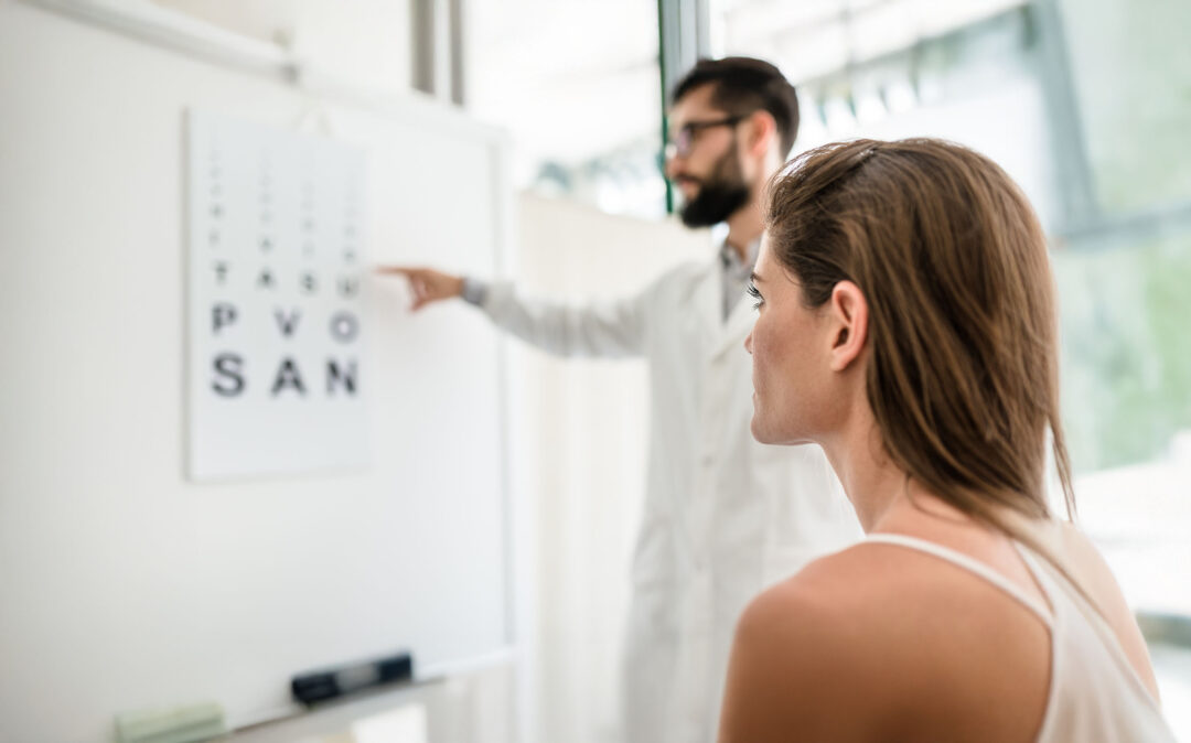 Eine Frau sitzt während eines Sehtests in einem hellen Untersuchungsraum, während ein Augenarzt auf eine Sehtafel an der Wand zeigt.