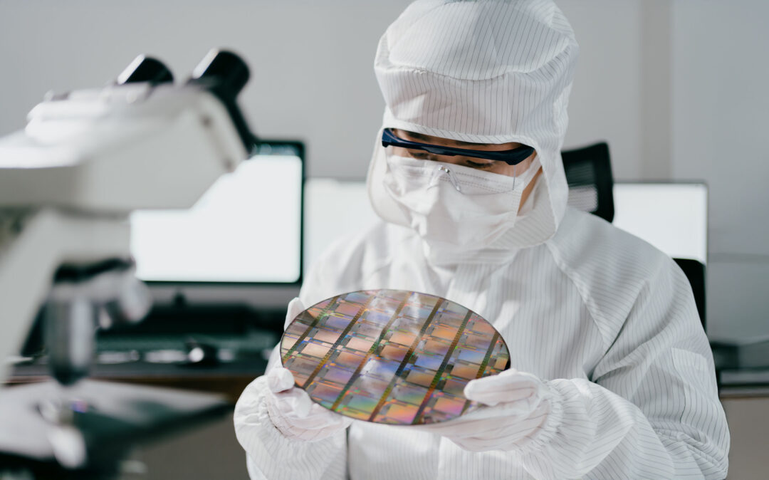 Ein Wissenschaftler in Reinraumkleidung untersucht einen Siliziumwafer in der Nähe eines Mikroskops in einer Laborumgebung.