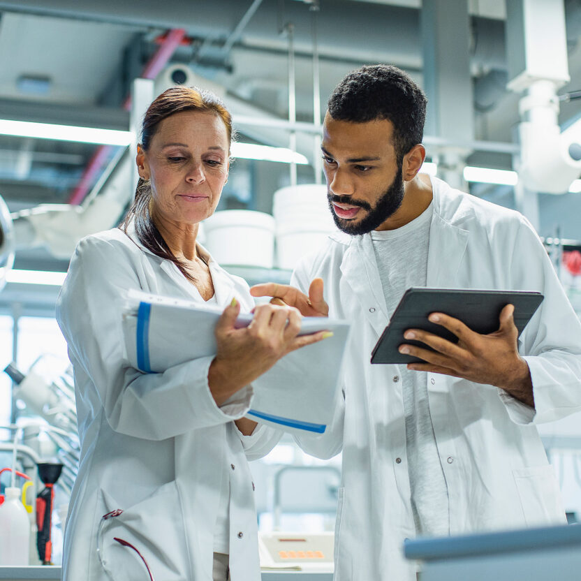 Zwei Wissenschaftler in Laborkitteln prüfen Notizen und Daten auf einem Klemmbrett und einem Tablet in einer Laborumgebung.
