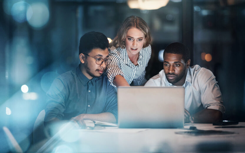 Drei Personen arbeiten nachts in einem Büro gemeinsam an einem Schreibtisch und schauen auf einen Laptop-Bildschirm.