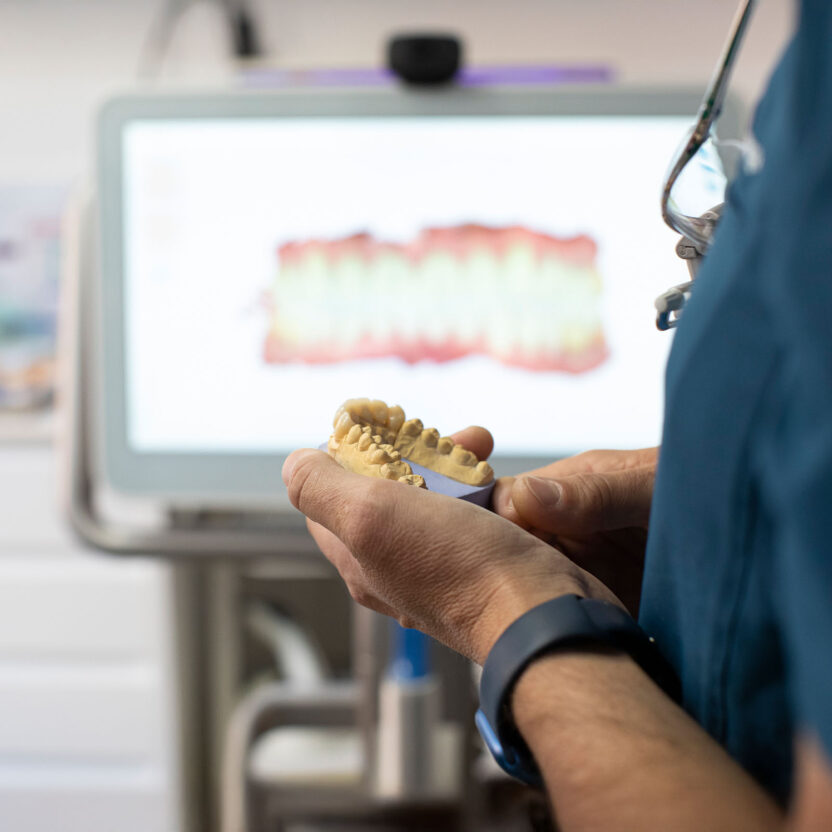 Eine Person im Kittel hält eine Zahnform vor einem Computer, der ein digitales Bild der Zähne in einer Zahnklinik anzeigt.