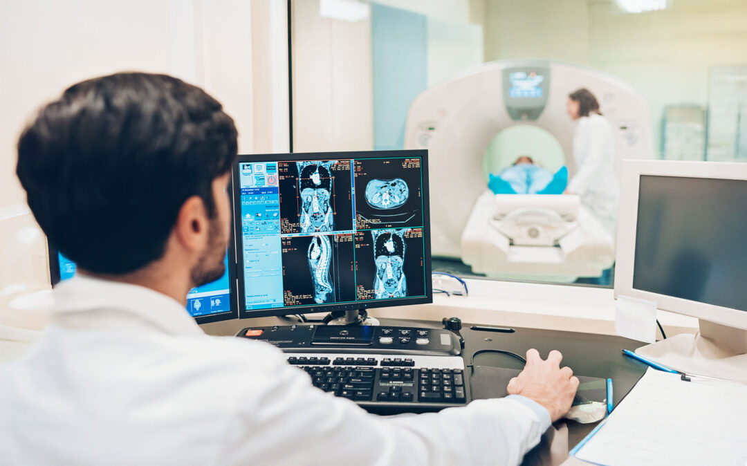 Ein Radiologe prüft MRT-Scans am Computer, während sich ein Patient im Nebenraum einem MRT-Verfahren unterzieht.