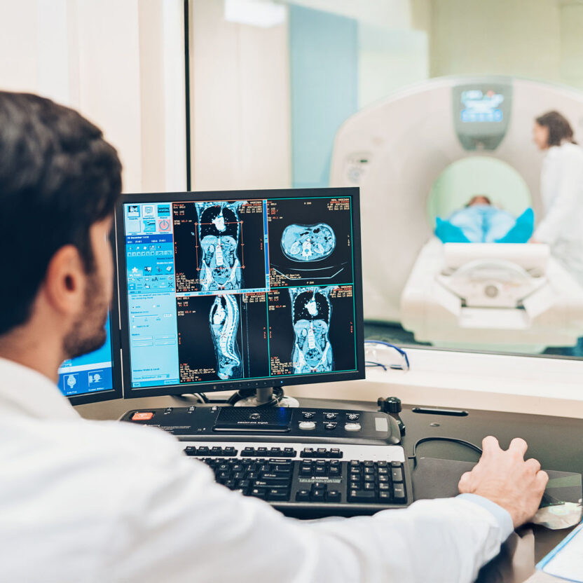 Ein Radiologe prüft MRT-Scans am Computer, während sich ein Patient im Nebenraum einem MRT-Verfahren unterzieht.