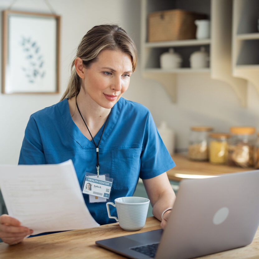 Eine Frau im Kittel sitzt an einem Küchentisch, hält Papiere in der Hand und schaut auf einen Laptop, wobei eine Tasse und ein Namensschild sichtbar sind.