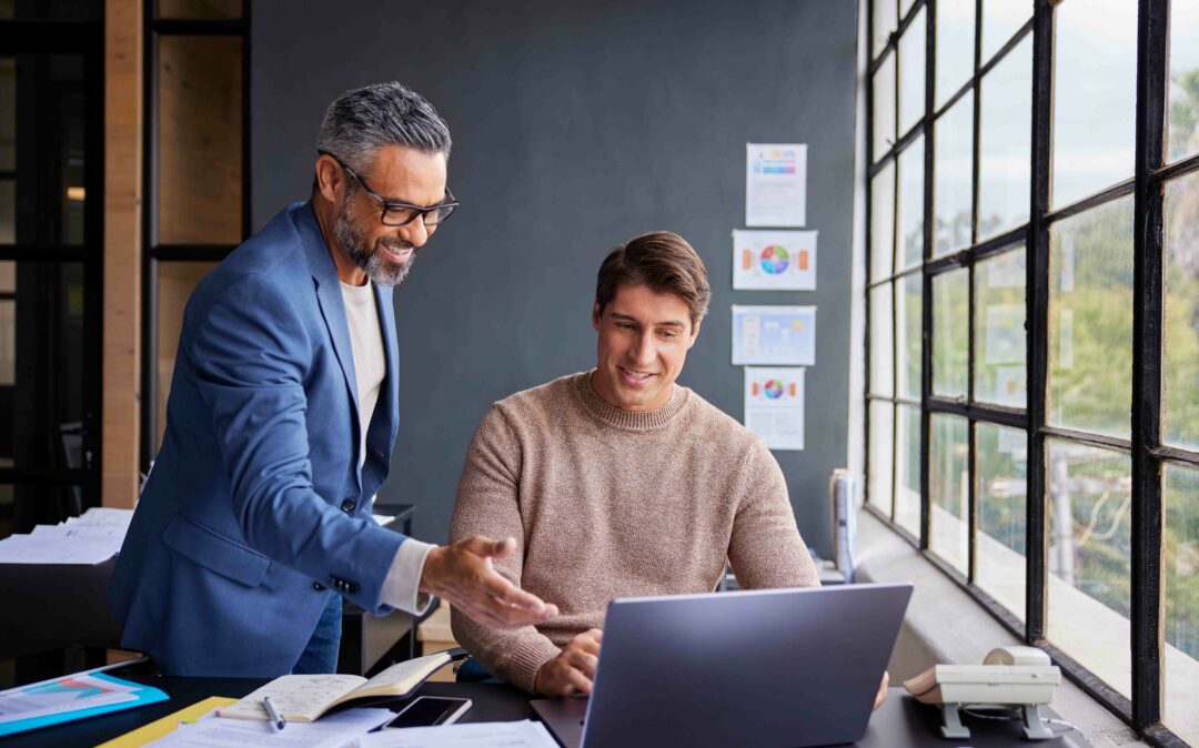 Zwei Männer in einem Büro schauen auf einen Laptop-Bildschirm, einer steht und gestikuliert, der andere sitzt und lächelt. Im Hintergrund sind große Fenster und Dokumente zu sehen.