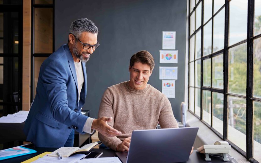 Zwei Männer in einem Büro schauen auf einen Laptop-Bildschirm, einer steht und gestikuliert, der andere sitzt und lächelt. Im Hintergrund sind große Fenster und Dokumente zu sehen.