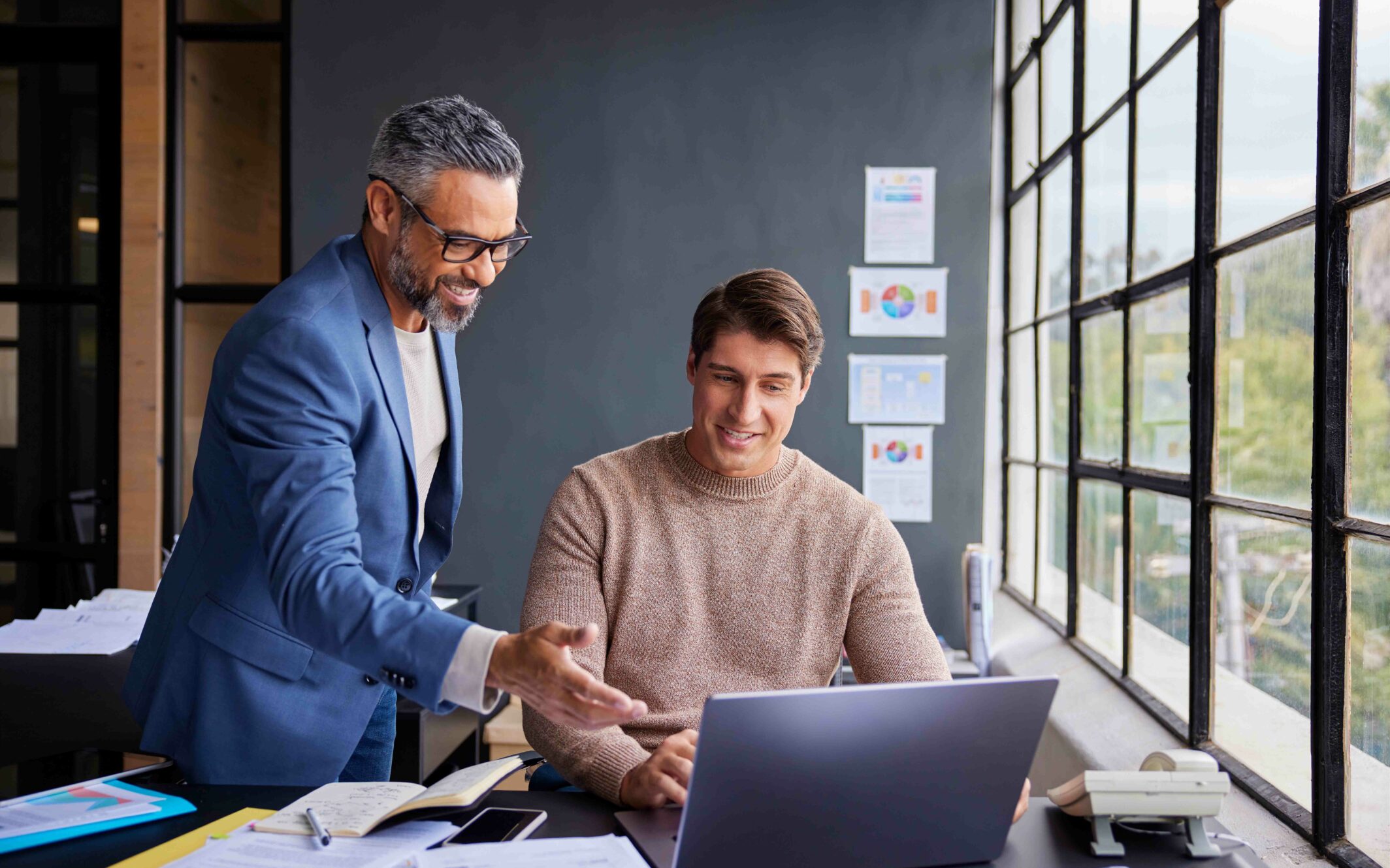 Two men in an office are looking at a laptop screen; one is standing and talking about SAP BNAC.