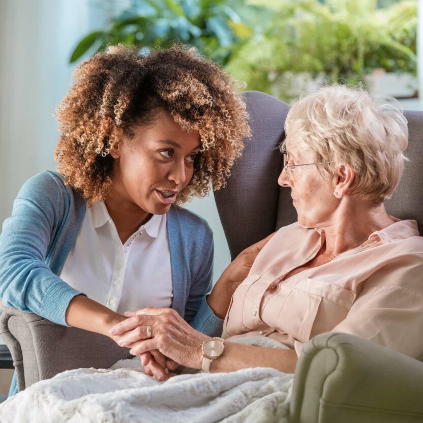 Eine jüngere Frau sitzt neben einer älteren Frau in einem Sessel, hält ihre Hand und spricht mit ihr in einem hellen, gemütlichen Raum mit Pflanzen.