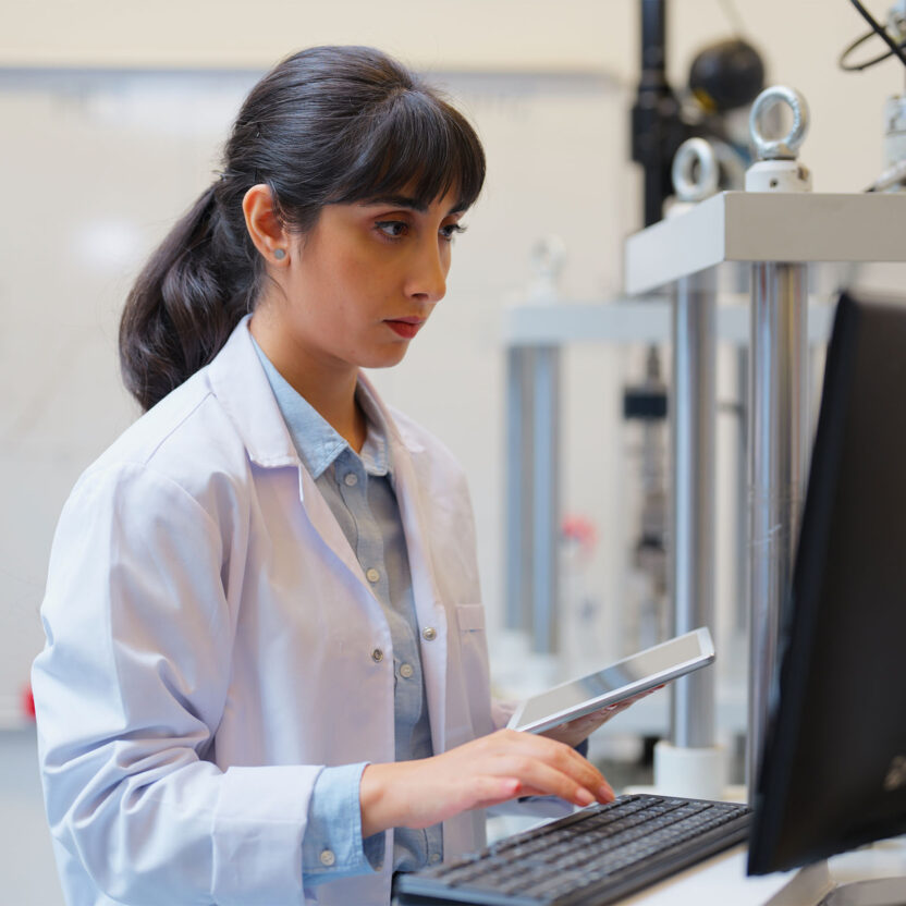 Eine Frau in einem Laborkittel benutzt eine Tastatur und ein Tablet, während sie an einem Computer in einer Laborumgebung arbeitet.
