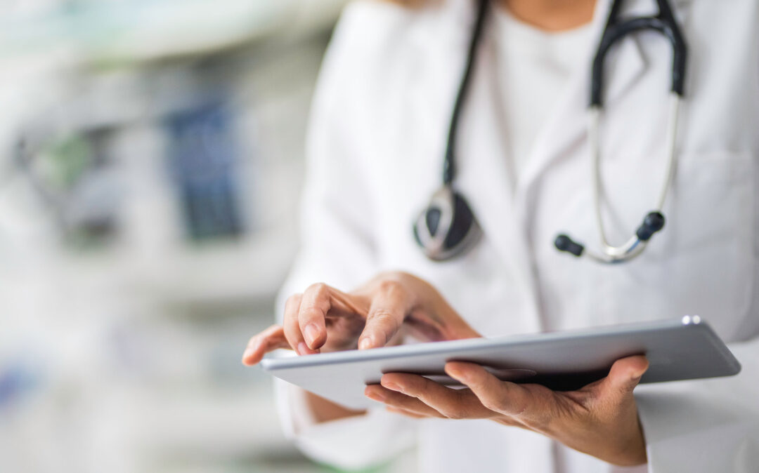 Ein Arzt im weißen Kittel mit Stethoskop benutzt ein Tablet, wobei der Hintergrund unscharf ist.