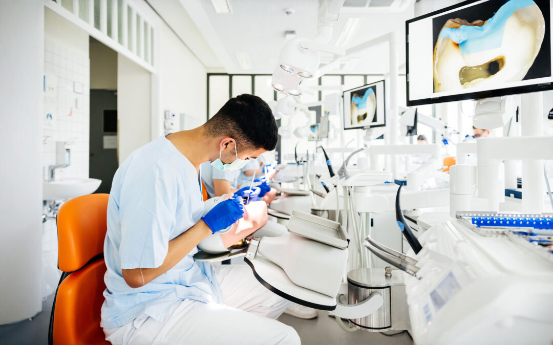 Ein Zahnarzt mit Handschuhen und Mundschutz behandelt einen Patienten in einer modernen Zahnklinik, wobei zahnärztliche Werkzeuge und ein Zahnbild auf einem Monitor zu sehen sind.