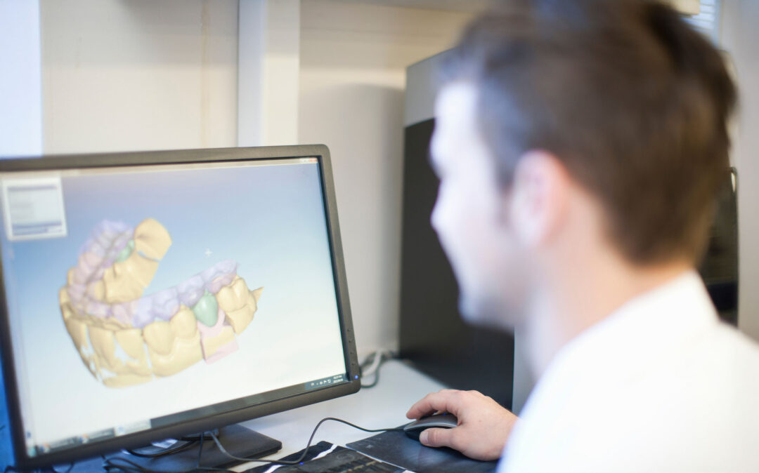 Eine Person sitzt an einem Schreibtisch und betrachtet ein 3D-Zahnmodell auf einem Computerbildschirm, was auf eine Tätigkeit in der Zahnmedizin oder Kieferorthopädie hindeutet.