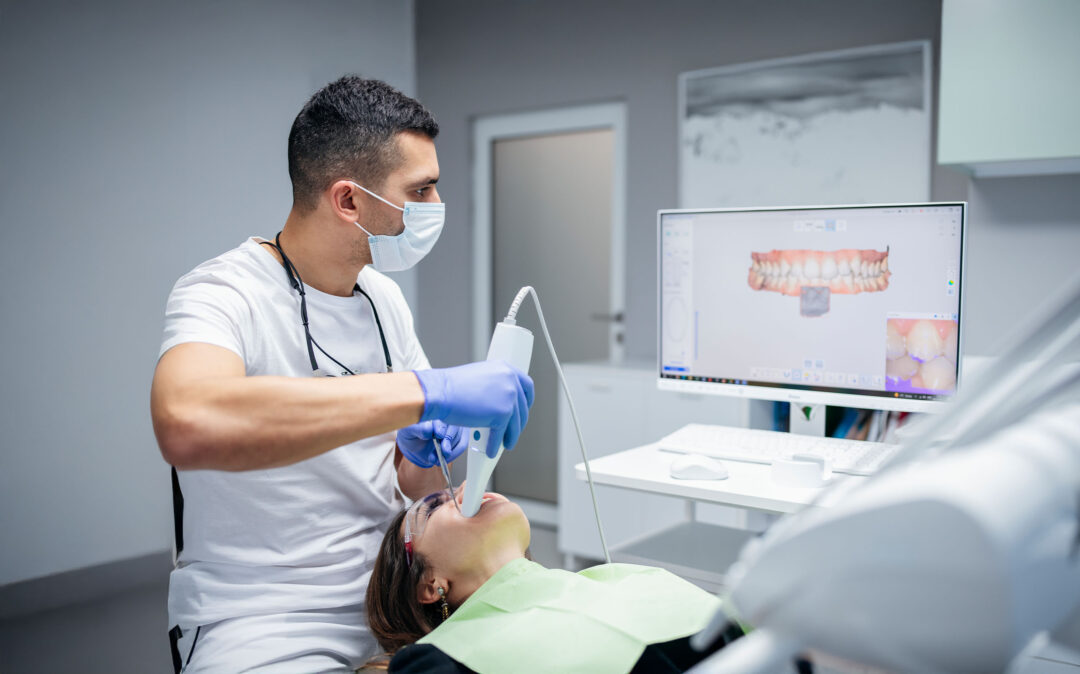 Ein Zahnarzt, der eine Maske und Handschuhe trägt, benutzt einen Handscanner im Mund eines Patienten, während ein Zahnscan auf einem Computerbildschirm in der Nähe angezeigt wird.