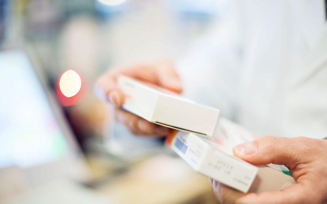 Eine Person in einem weißen Kittel hält zwei rechteckige Arzneimittelboxen in den Händen, deren Hintergrund unscharf und ein rotes Licht sichtbar ist.