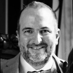 A man with short hair and a beard, wearing a suit and bow tie, smiles at the camera in a black-and-white portrait.