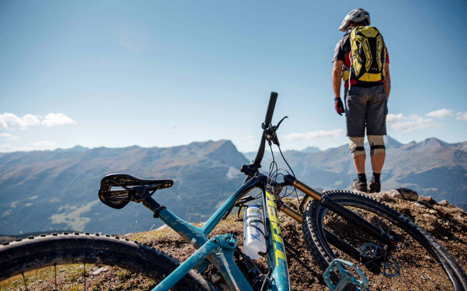 Bild von einem Mountainbike-Fahrer, der sein Fahrrad im vorderen Bereich des Bildes abgelegt hat & sich auf einen Felsen gestellt hat, von wo er ins Gebirge blickt.
