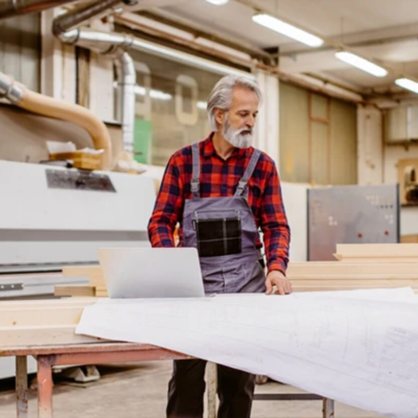 Handwerker mit grauem Bart betrachtet Baupläne in einer Werkstatt.