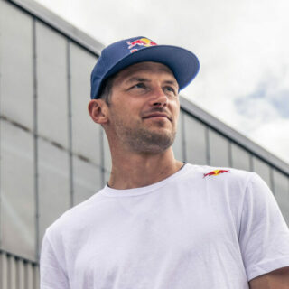 Peter Salzmann wearing a white Red Bull T-shirt and blue cap, looking into the distance in front of a modern building under a cloudy sky, photo by Mirja Geh.
