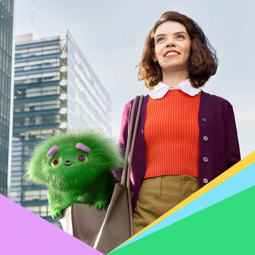 Junge Frau lächelt selbstbewusst vor modernen Bürogebäuden, während ein grünes, plüschiges Fantasiewesen aus ihrer Tasche schaut – Symbolbild für karriere.at und einen positiven Start ins Berufsleben.