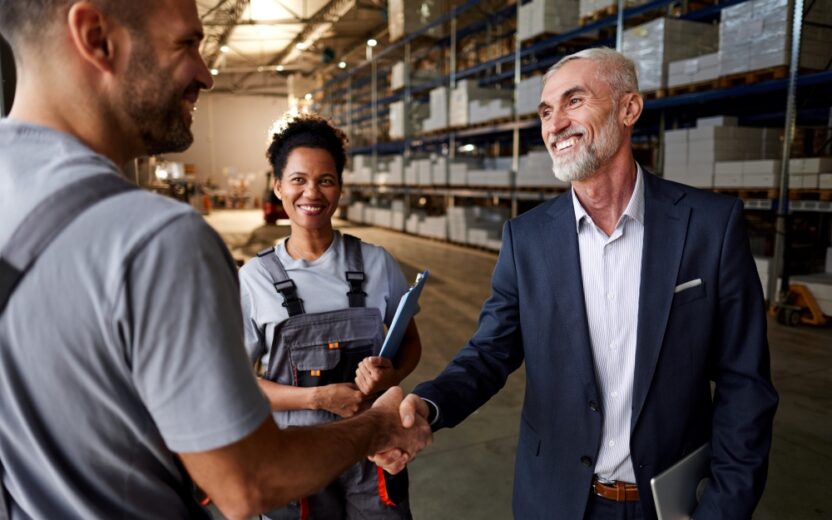 Happy manager and manual worker shaking hands next to female colleague in distribution warehouse