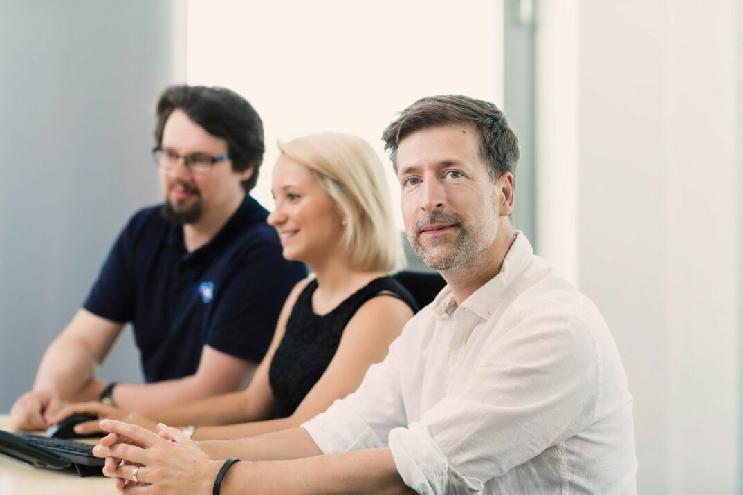 Drei Personen sitzen an einem Tisch, ein Mann schaut direkt in die Kamera.