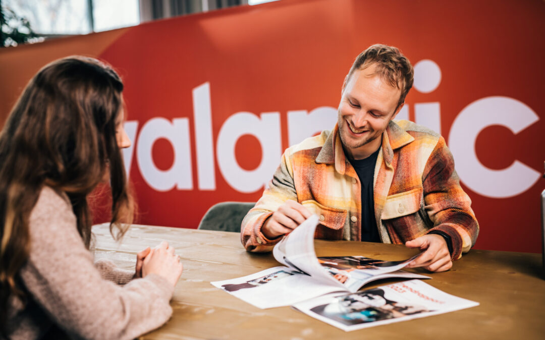 Two people sit at a table, one showing the other a magazine. Both are engaged in conversation, with a red wall and partial text "alamic" in the background.