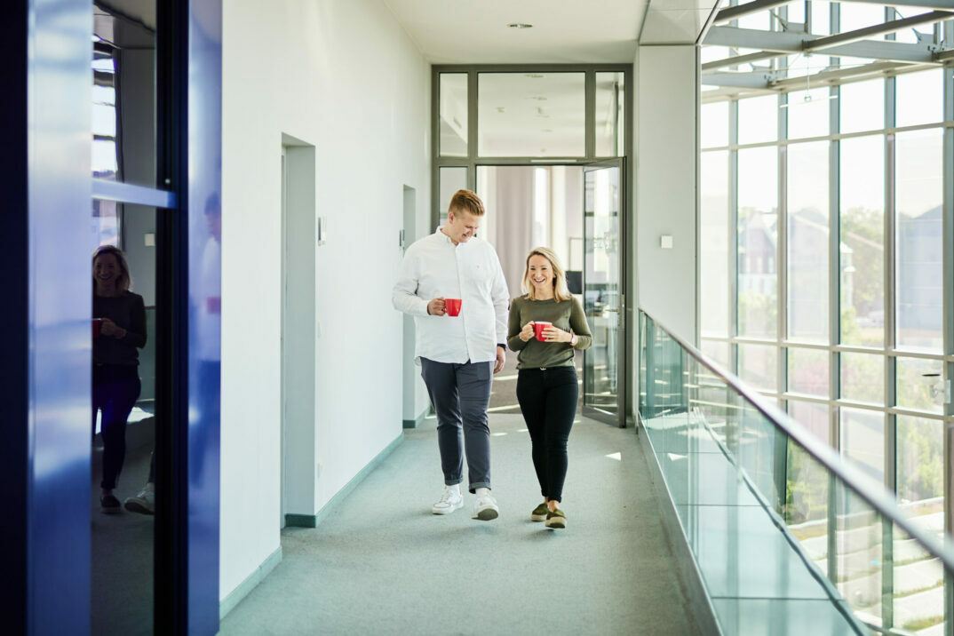 Photo of a man and a woman walking down an office hallway, each holding an orange coffee cup.