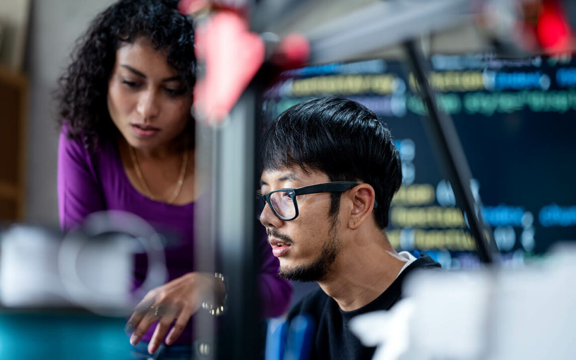 Two people are concentrating on a computer screen, behind which there is a code.