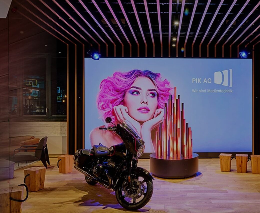 Vor einem großen Bildschirm mit "PIK Ag" ist ein Motorrad abgestellt.