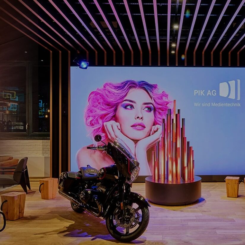 Vor einem großen Bildschirm mit "PIK Ag" ist ein Motorrad abgestellt.