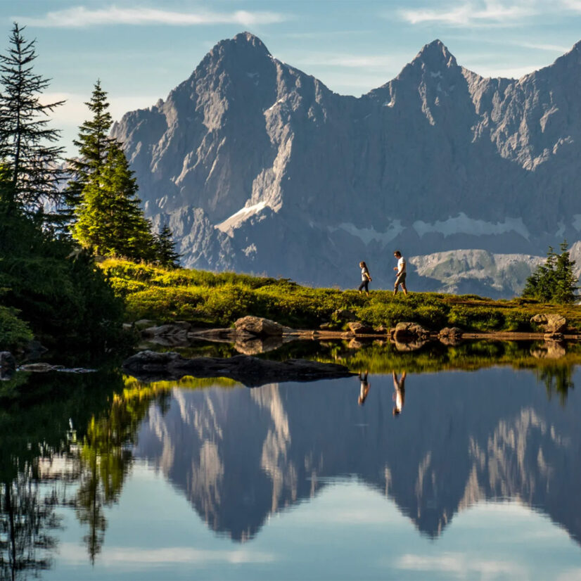 Ein Wanderer mit seinem Kind spaziert an einem sonnigen Tag entlang des Spiegelsees in der Steiermark. Im Hintergrund sieht man herrliches Bergparomana.