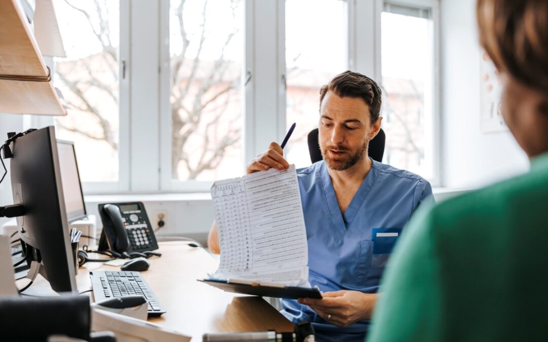 Männlicher Arzt liest Krankenakte eines Patienten, während er im Untersuchungszimmer sitzt.
